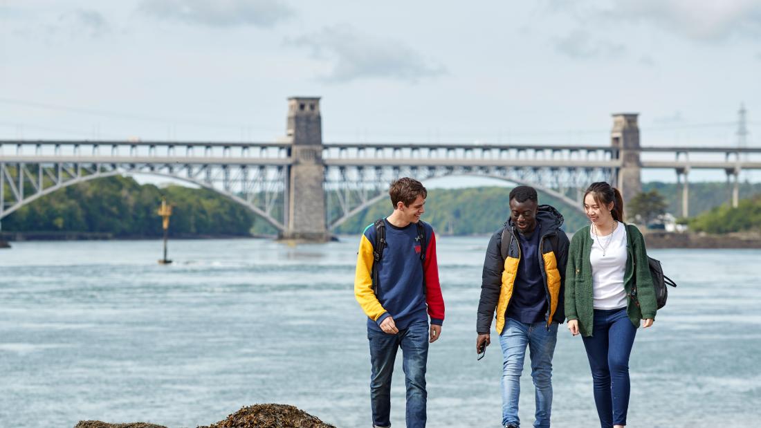 Three students walking along the beach with the Britannia bridge in the background
