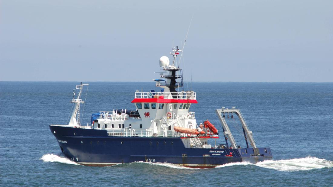 A blue and white research vessel creates a bow wave in a blue sea