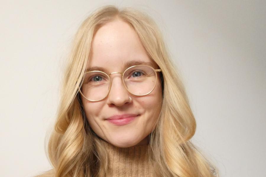 Young woman with long blond hair and light colour-framed spectacles smiles into camera.