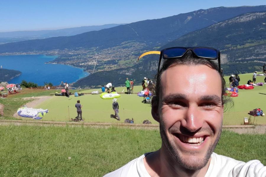 Marley Willegers smiles into camera with sunglasses onhis head, behind him are people preparing to paraglide and  landscape with lake far below and beyond.