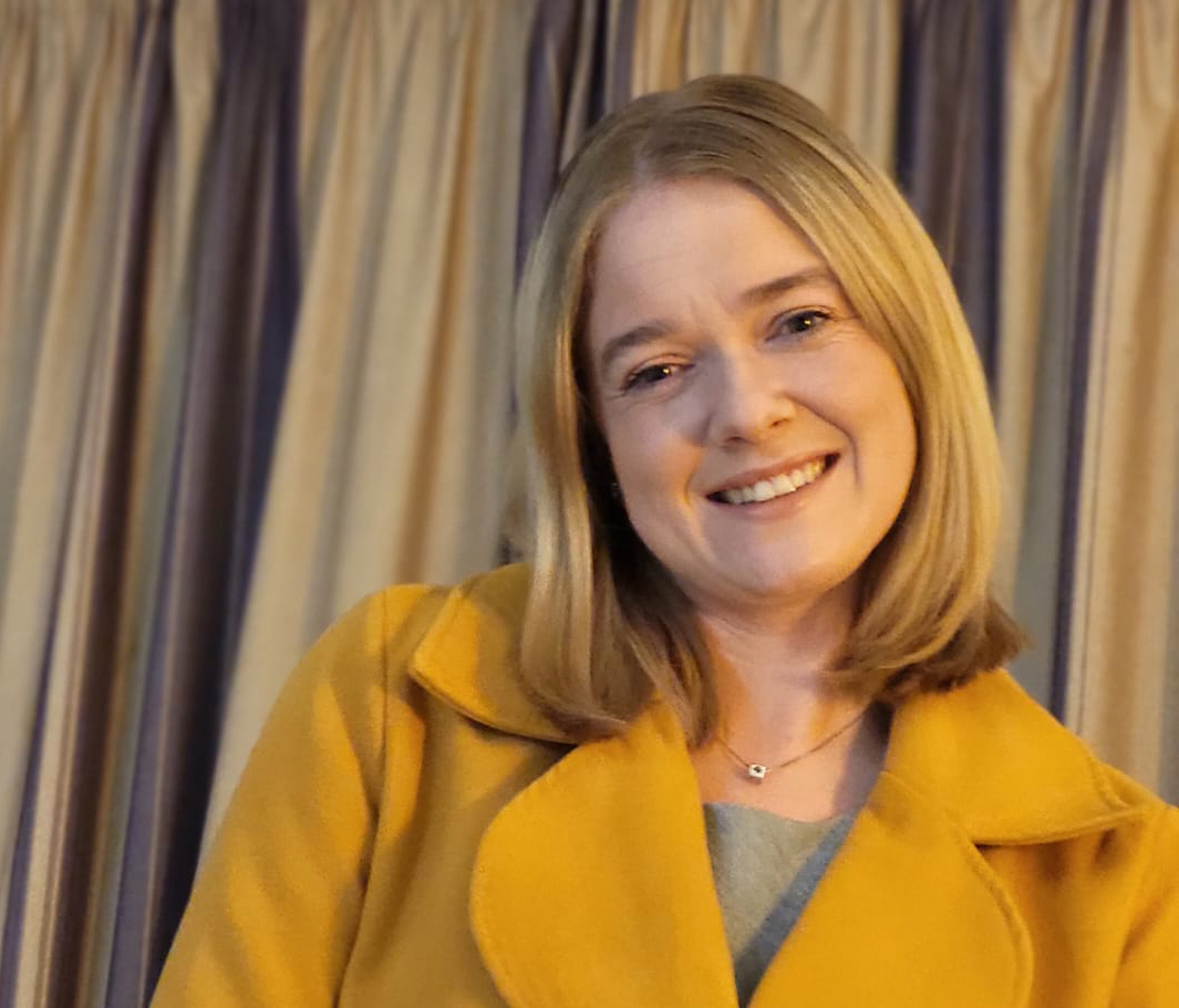 Megan Phillips smiling to camera in orange coat with curtain behind her