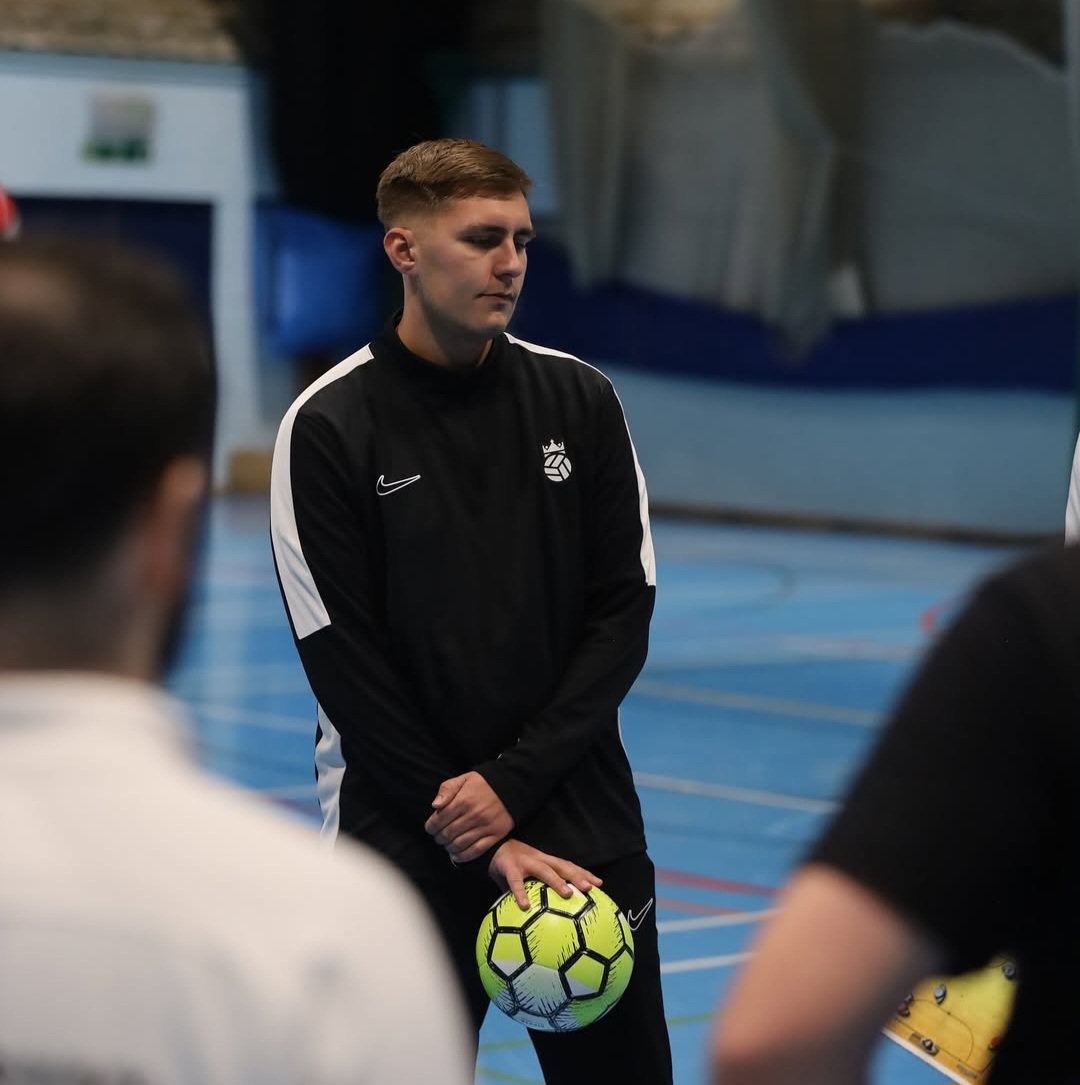 Glenn Swain standing on indoor football pitch in black kit with a yellow football
