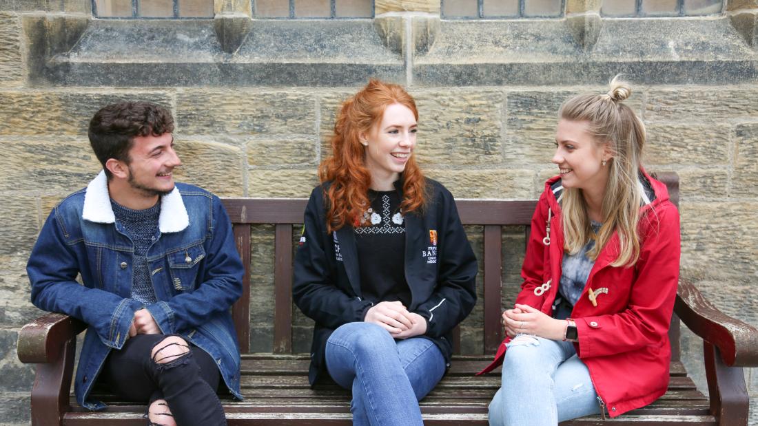 Three students sat on a bench on campus talking to each other 