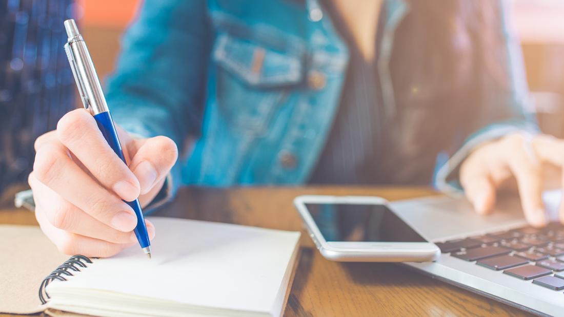 Close-up of student writing in notepad with mobile phone and laptop on desk