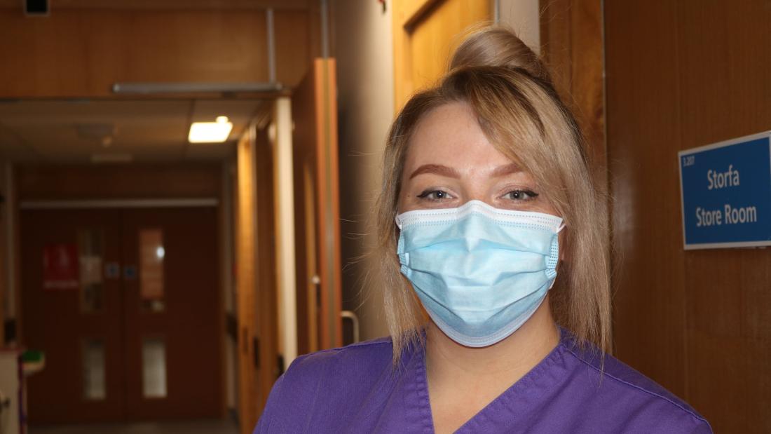 A student nurse wearing  purple uniform and a mask looks into the camera