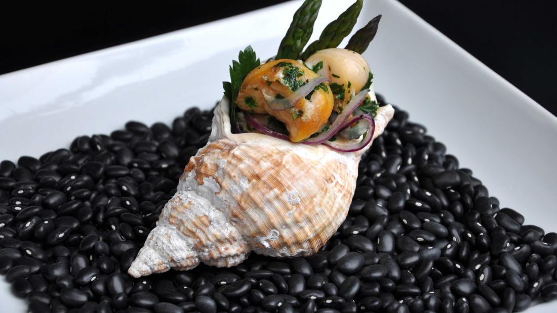 a cooked whelk on a bed of black beans on a square white plate