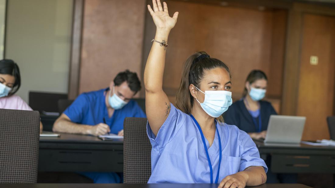 A woman in 'scrubs' and mask holds up her hand in a classroom, others are writing