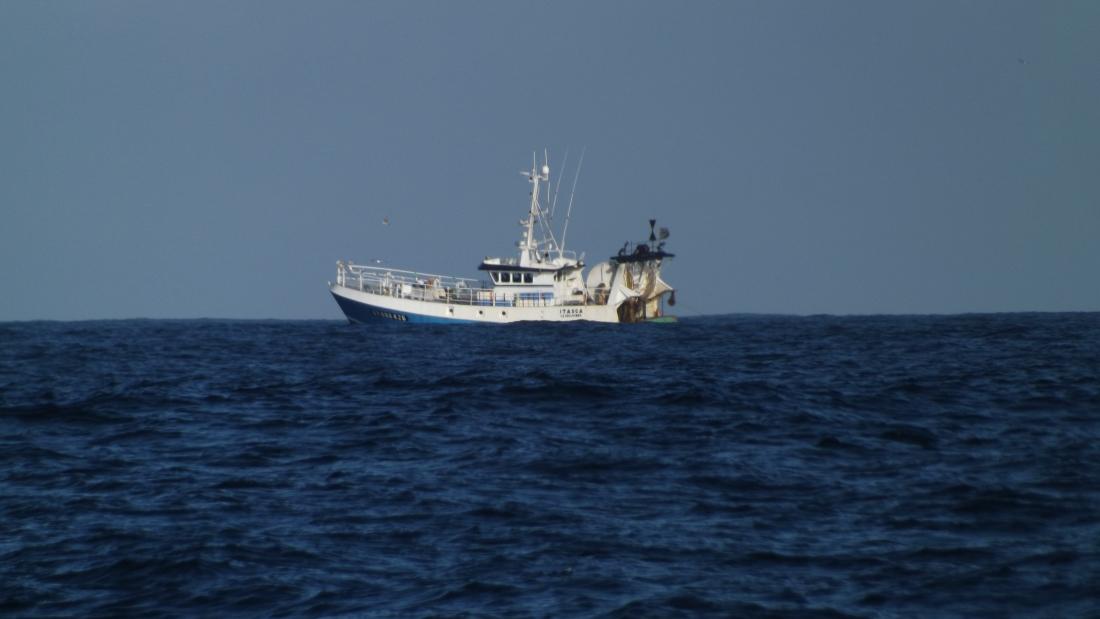 A blue trawler sits near the horizon on a blue sea under a lighter blue sky.