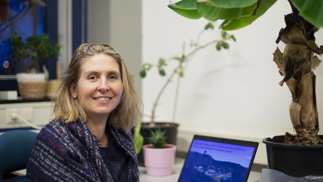 Professot Julia Jones, with blonde shoulder length hair smiles into camera, she is seated in front of a laptop computer and large tropical looking houseplant.