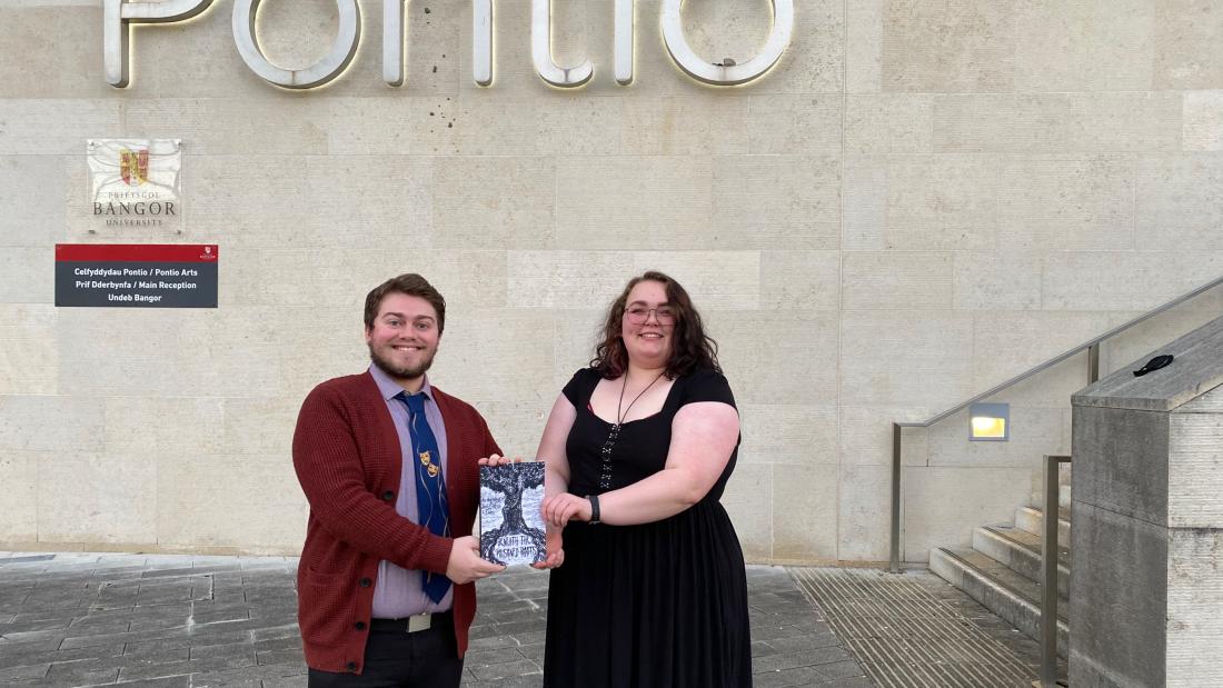 Dau fyfyriwr o Brifysgol Bangor yn dangos eu cyhoeddiad cyntaf y tu allan i adeilad Pontionts show the book they have published while standing in front f the external of the Pontio building