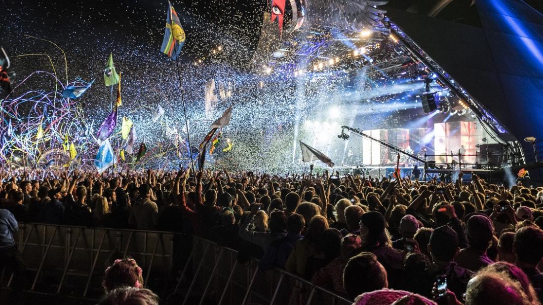 A large crowd looks towards a brightly lit stage at night. Above their heads are coloured banners.