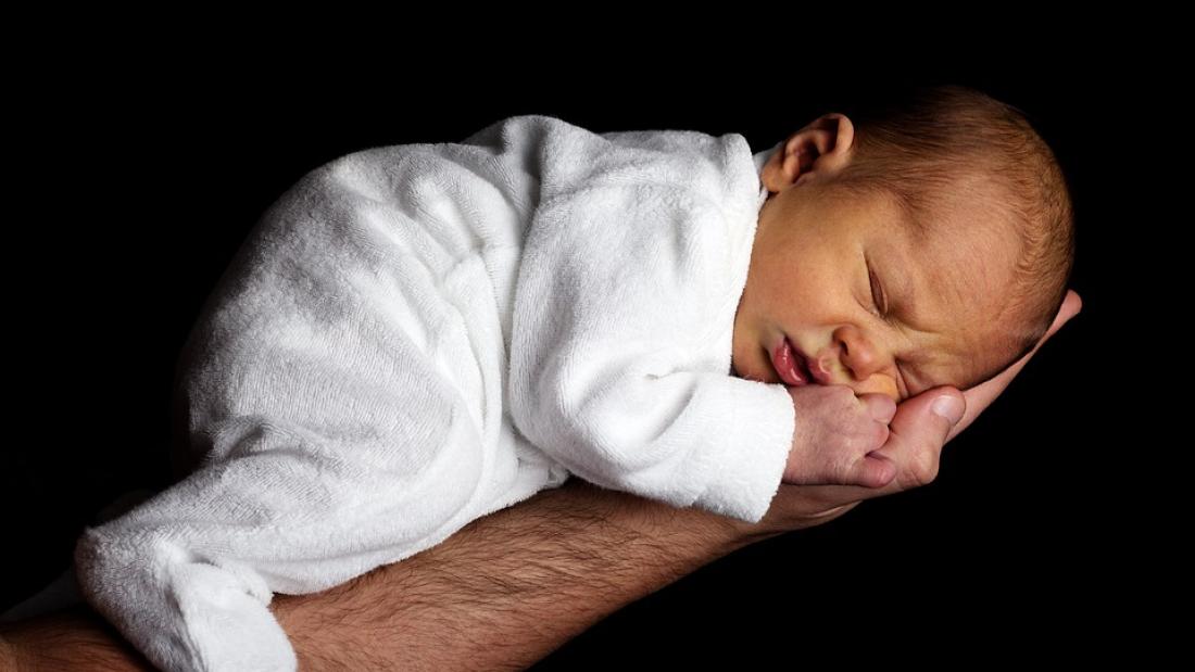A small baby in a white sleepsuit sleeps in a man's hand and forearm.