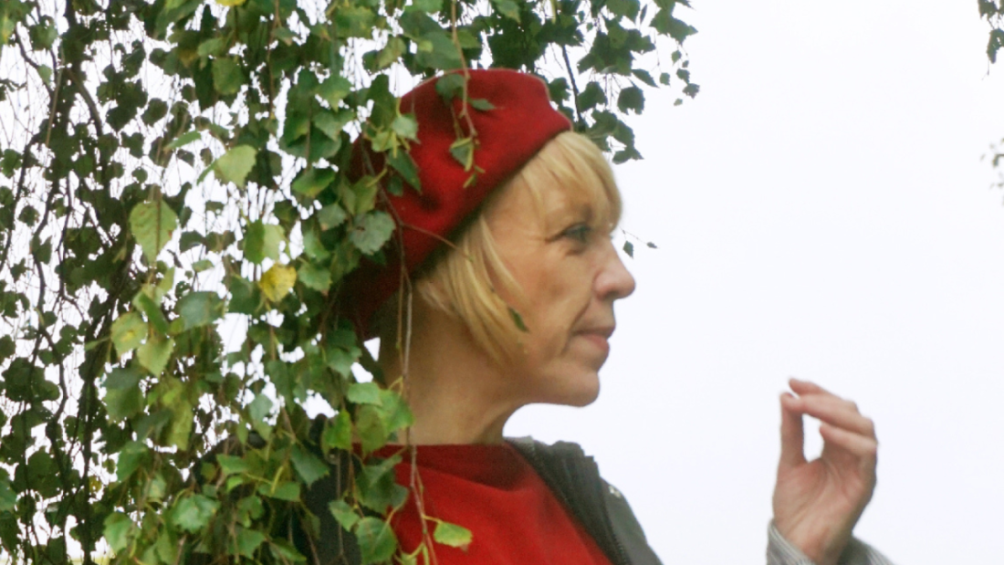 Woman with blonde hair wearing red and a red beret stands directly under a tree in leaf.