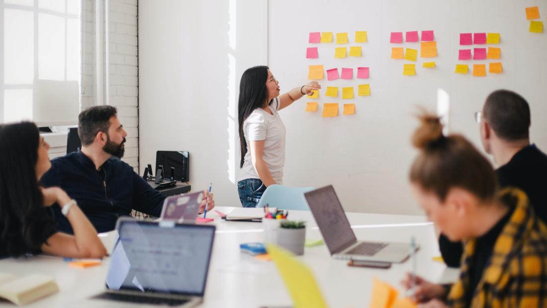 Woman in front of board with sticky notes - room of people- training session
