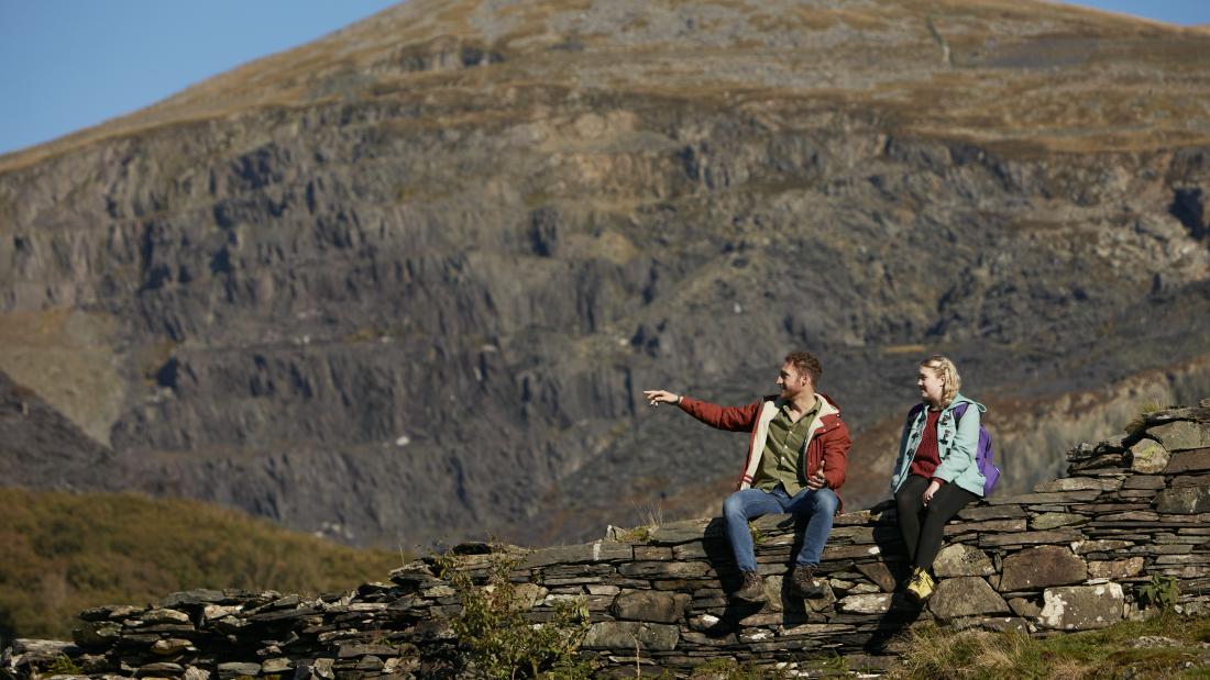 Young aman and woman sit on a slate wall, in the background is an enormous slate quarry in the side of a mountain