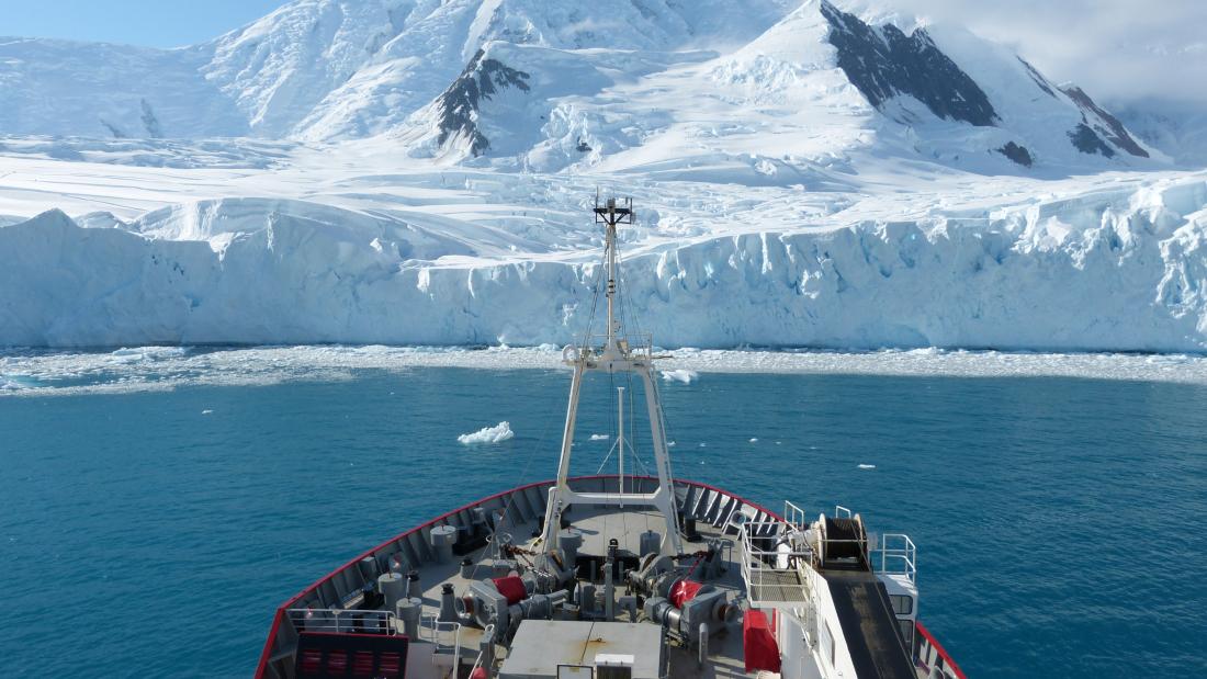 The bow of a ship is seen in front of snow covered mountains and a glacier running down to a blue sea.