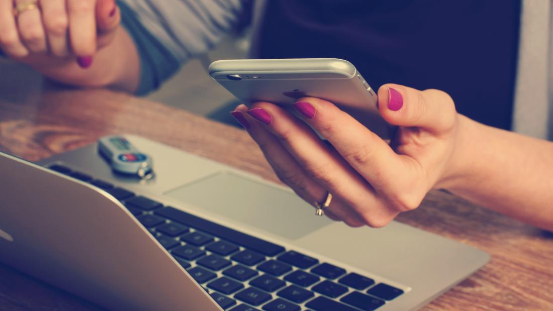 An image of someone working in front of a laptop holding their mobile phone