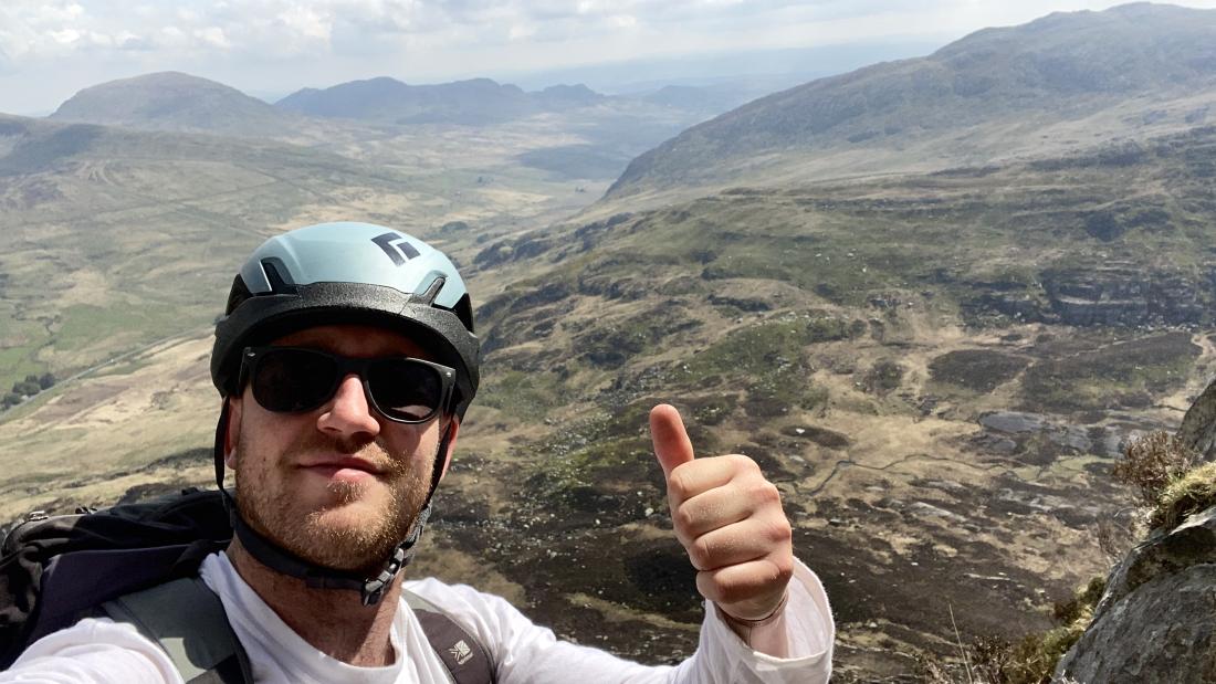 George, wearing sunglasses and climbing helmet gives a thumbs up sign,  as he prepares for his Last Pole expedition. There are mountains in the background.