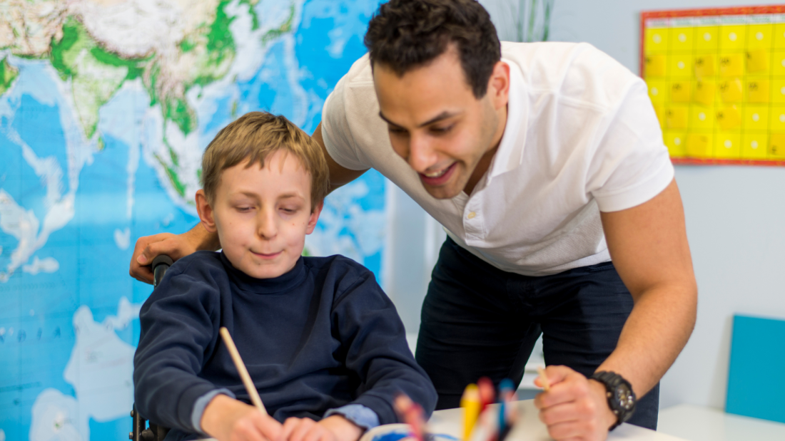 Young man standing with a younger male child who is sitting at a desk