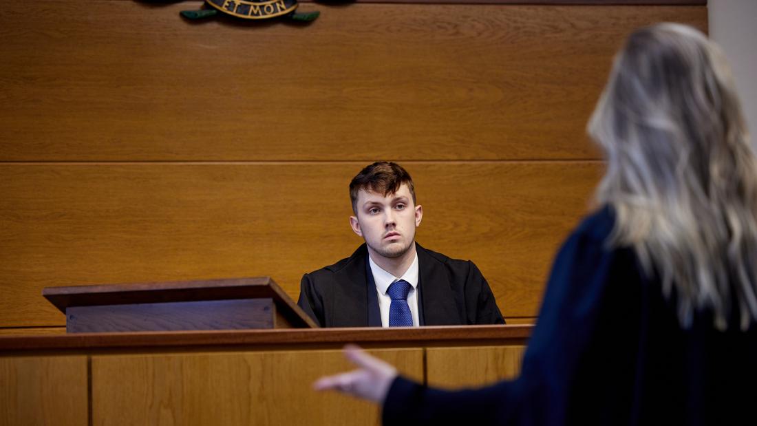 Student sitting behind desk whilst talking to other student practicing mooting in the mock moot court room 