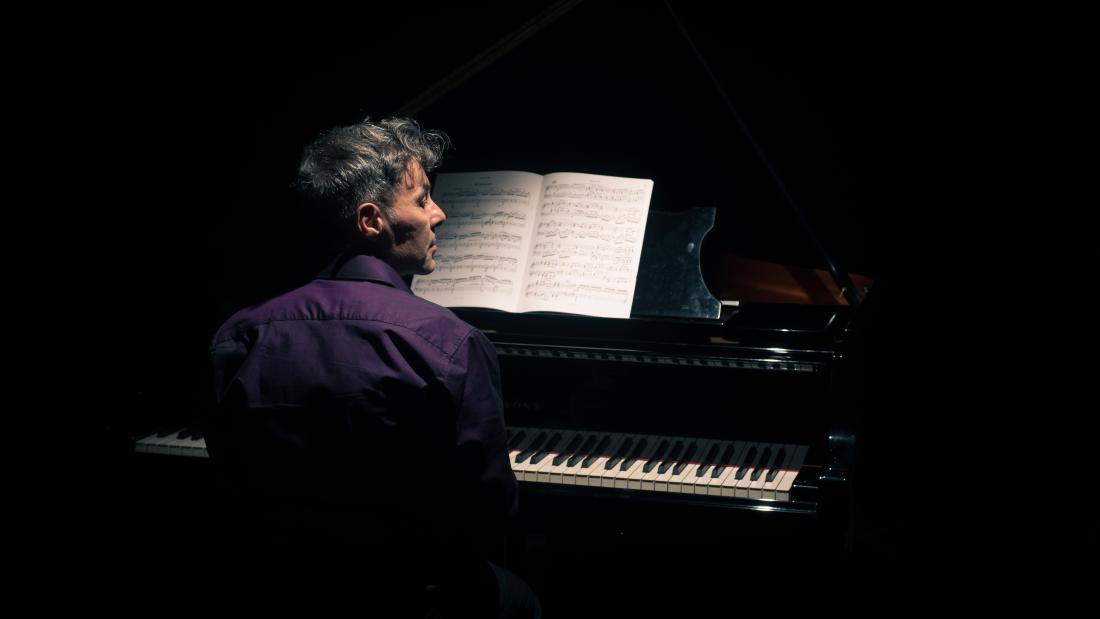 Man in purple shirt with short gerying hair plays piano and looks to the right, so we see his face in profile from the back, we see him and the piano keys the rest is black.