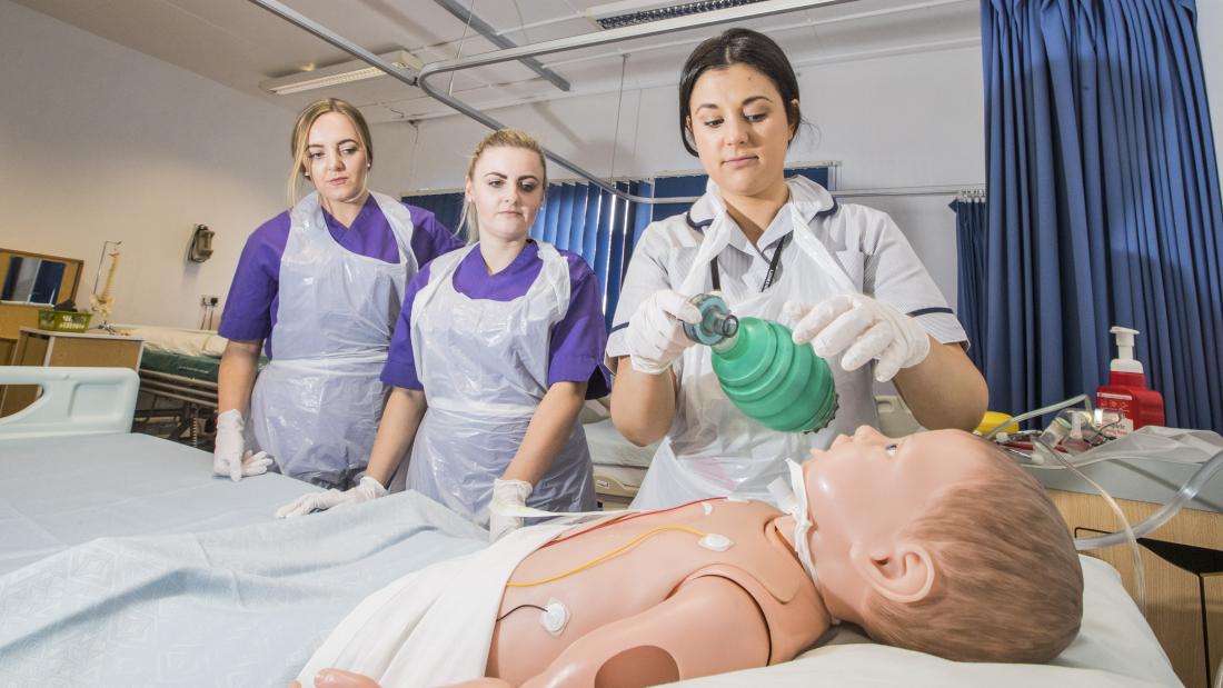 Two nursing students at a bedside watching someone with a dummy