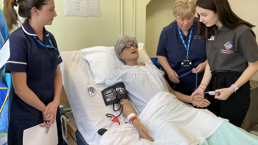 YOung girl approached a dummy in a bed- there are nurses on either side.