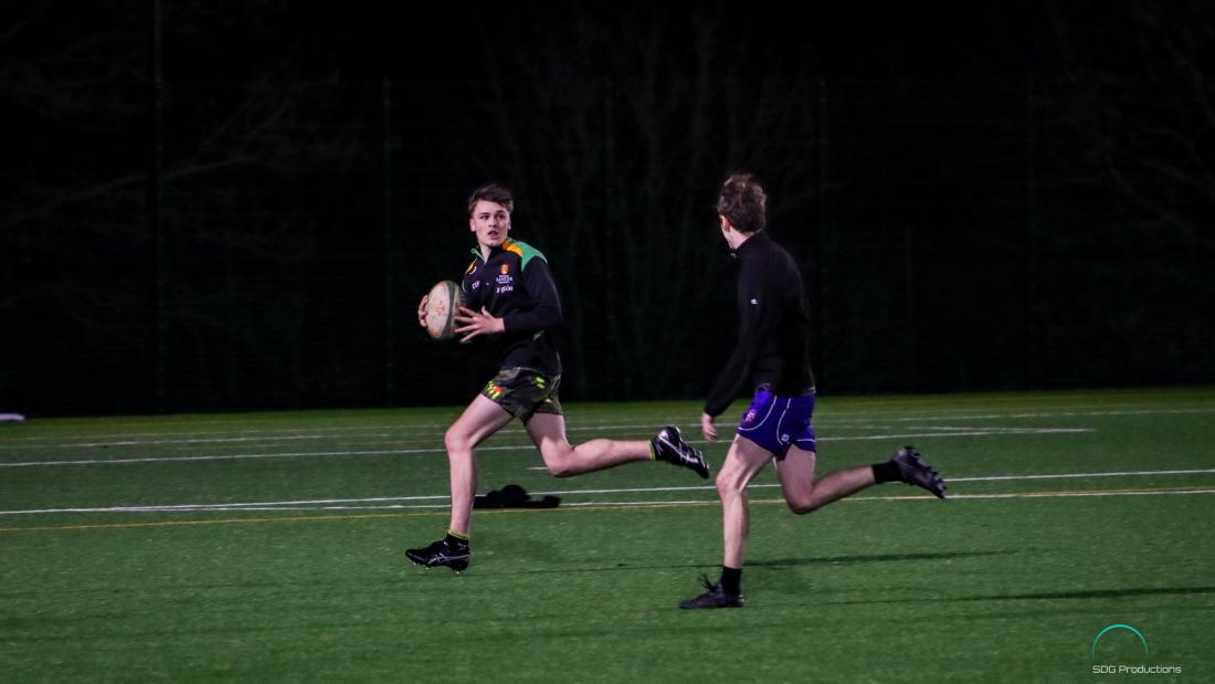 Two rugby players, one running with the ball looks back towards the other approaching from the side.