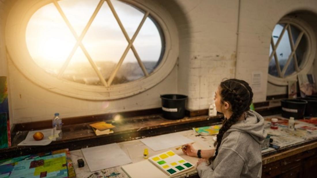 A student with plaits at a work desk, looks through a window