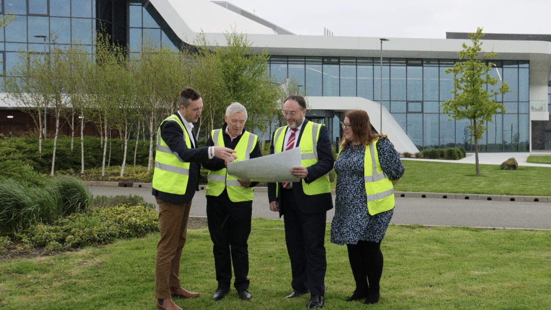  pedwar person mewn wasgod amlygu yn edrych ar gynllun tu alan i'r adeilad presennolplans outside the current building.