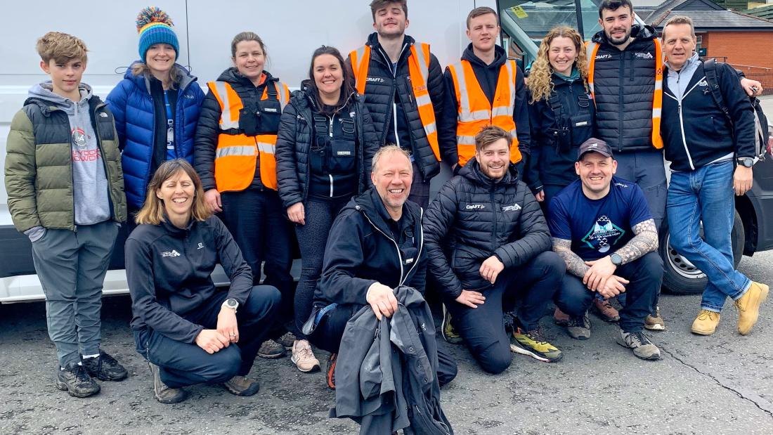 Group of men and women, some in hi viz jackets standing  and crouching in front of a van