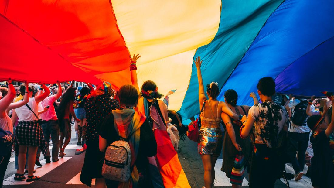 people parade under an enormous rainbow pride flag