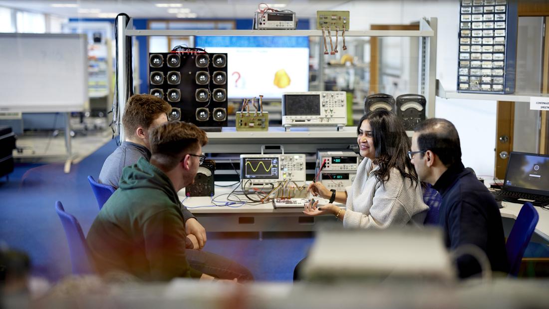 Four students sitting in a circle talking to each other, in an electronic engineering laboratory, with equipment in the background.