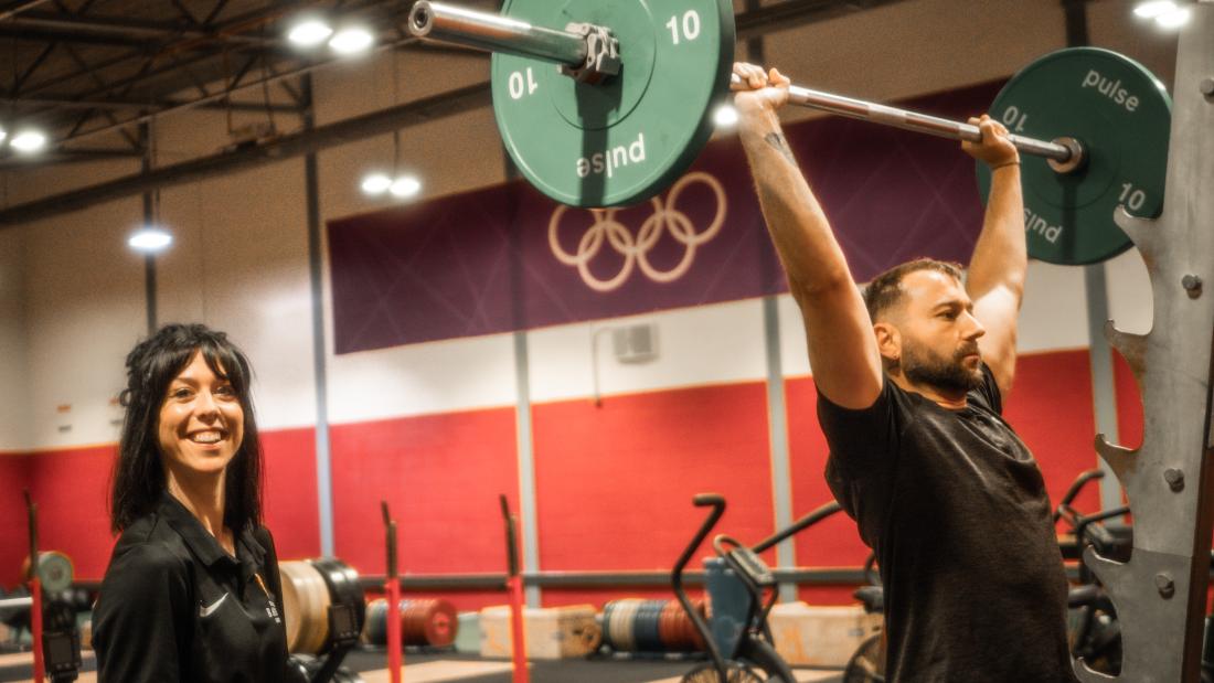 An instructor smiling at the camera while a client lifts weight