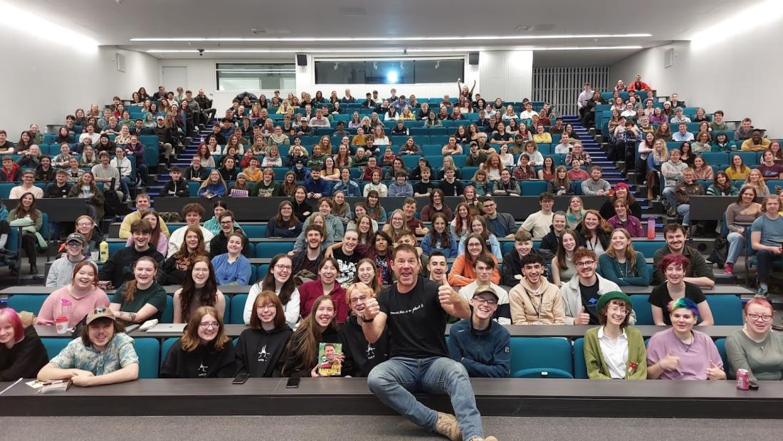 Steve Backshall yn chwifio'i fodiau o flaen darlithfa lawn