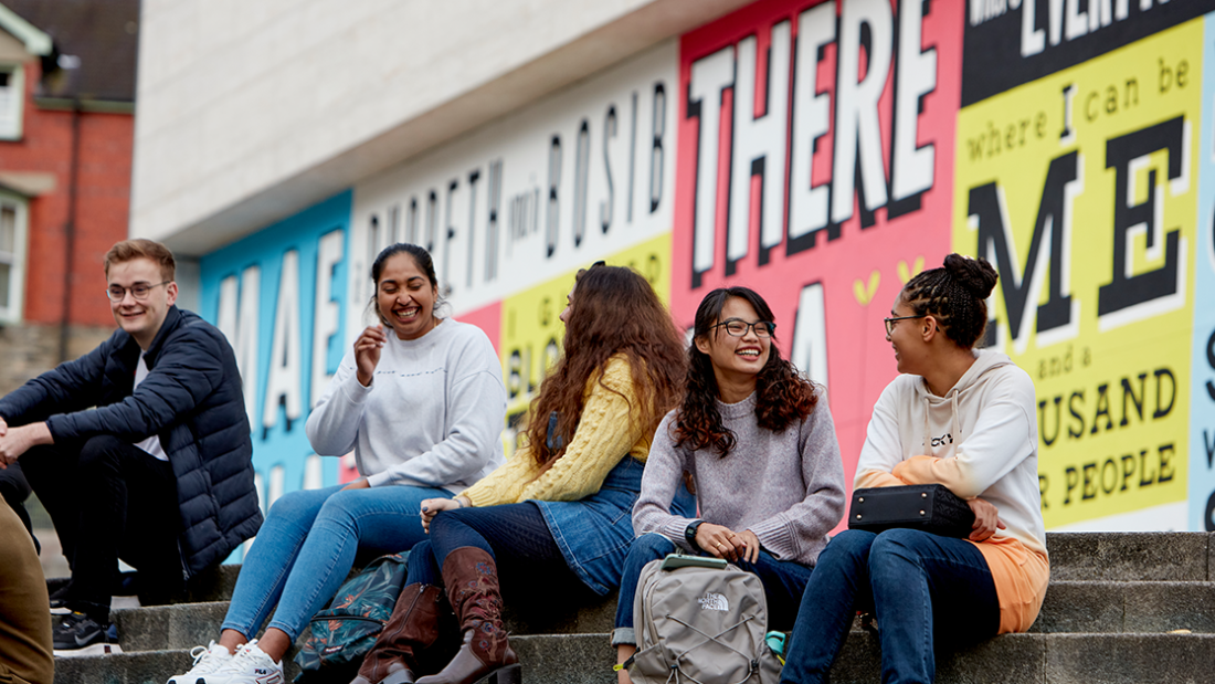 students sitting outside Pontio on the steps in front of a colourful mural.