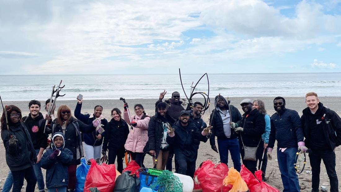 A group made up of International students, staff and children on a beach surrounded by litter bags having recently undertaken a beach clean up 