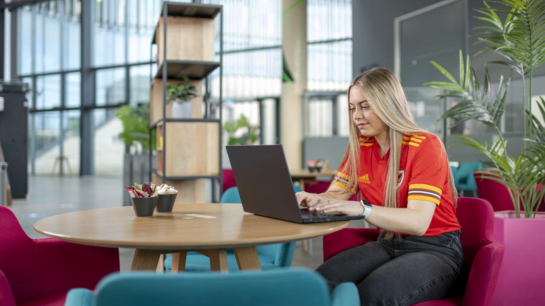 Myfyriwr yn eistedd yn gweithio ar liniadur yn Pontion yn gwisgo crys-t pêl droed Cymru