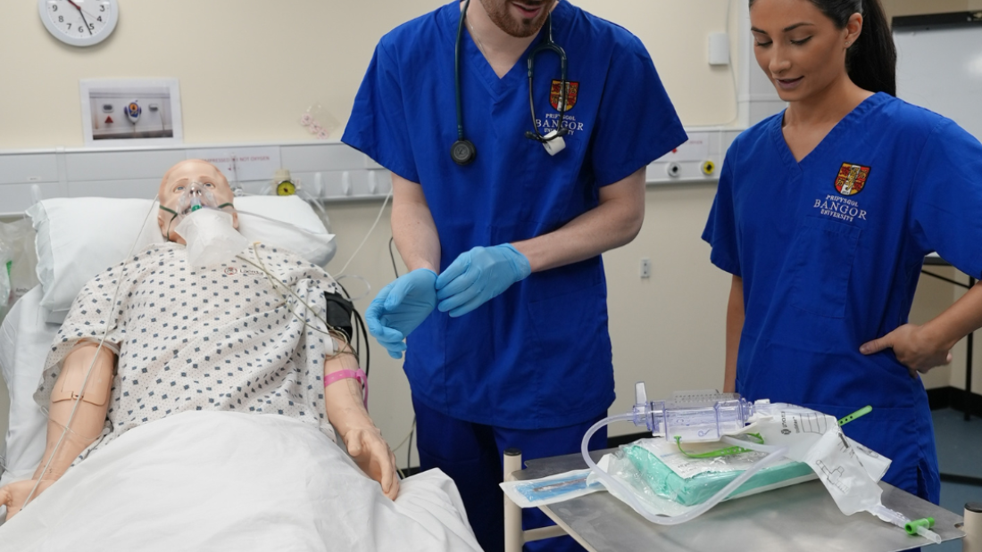 a simulation mannequin is lying in a hospital bed in a university clinical skills suite. two medical students a male and a female are stood to the side with a trolley
