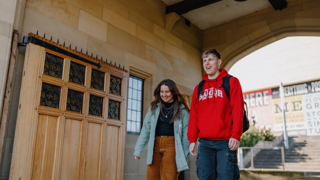 Two students walking through memorial arch one wearing a red Bangor University hoodie the other a blue coat