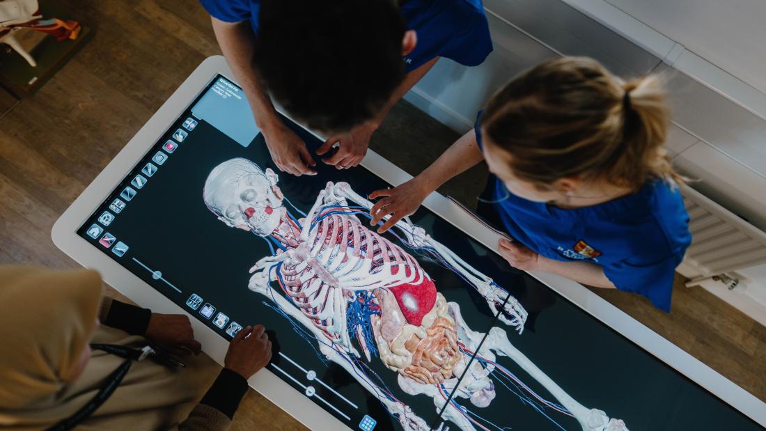 a human skeleton is shown on the screen of a digital anatomy table from above with 2 students and a lecturer stood around it