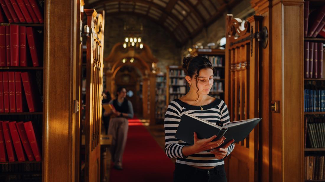 A student stands in the central aisle of Shankland Library reading a book, surrounded by bookshelves with warm light filtering through high, arched windows. The vaulted ceiling and chandeliers add a sense of quiet grandeur.