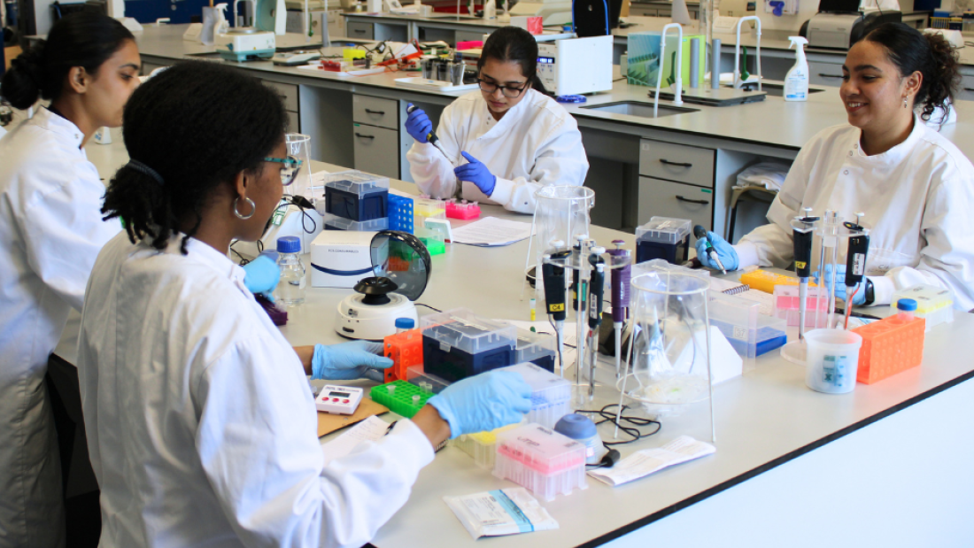 4 female students wearing lab coats are in a laboratory sat around a bench. There is an array of lab equipment on the bench and some students are using the equipment.