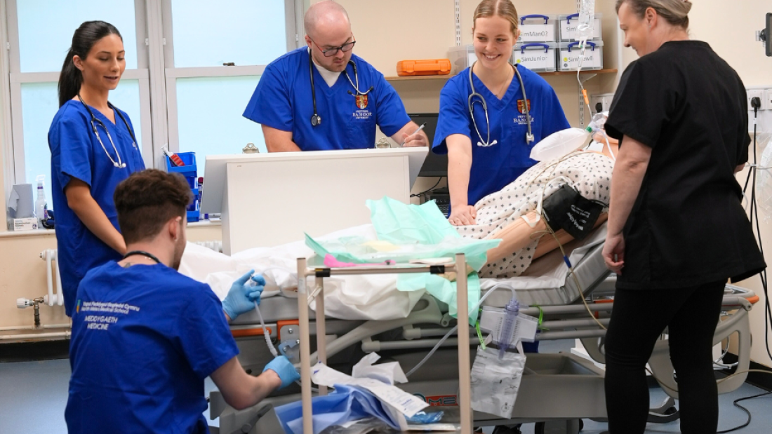 4 medical students dressed in blue scrubs stand around a mannikin in a hospital bed, there is an intructor in a black tunic overseeing the students