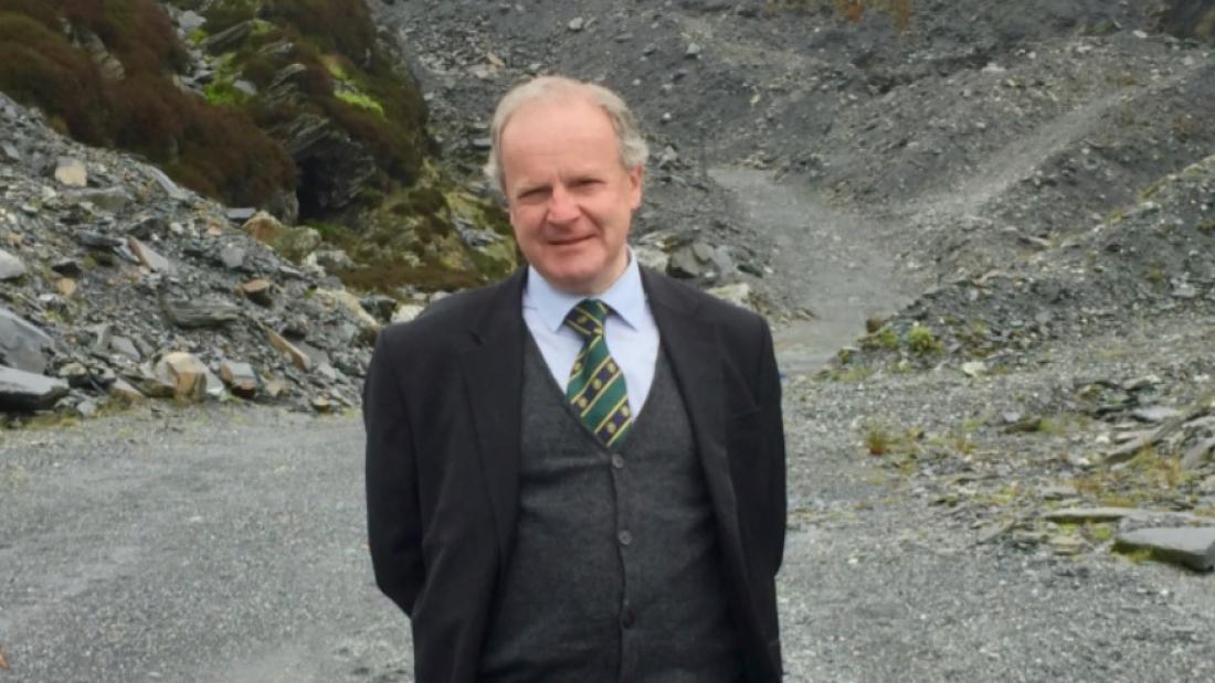 A photograph of David Gwyn smiling, behind him is an example of the slate landscape, with a path that is flanked by slate slag heaps 