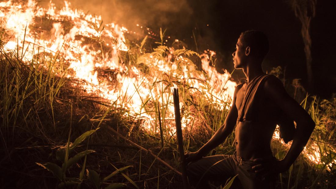 A man stood by some burning vegetation, he is lite by the bright orange flames of the fire.