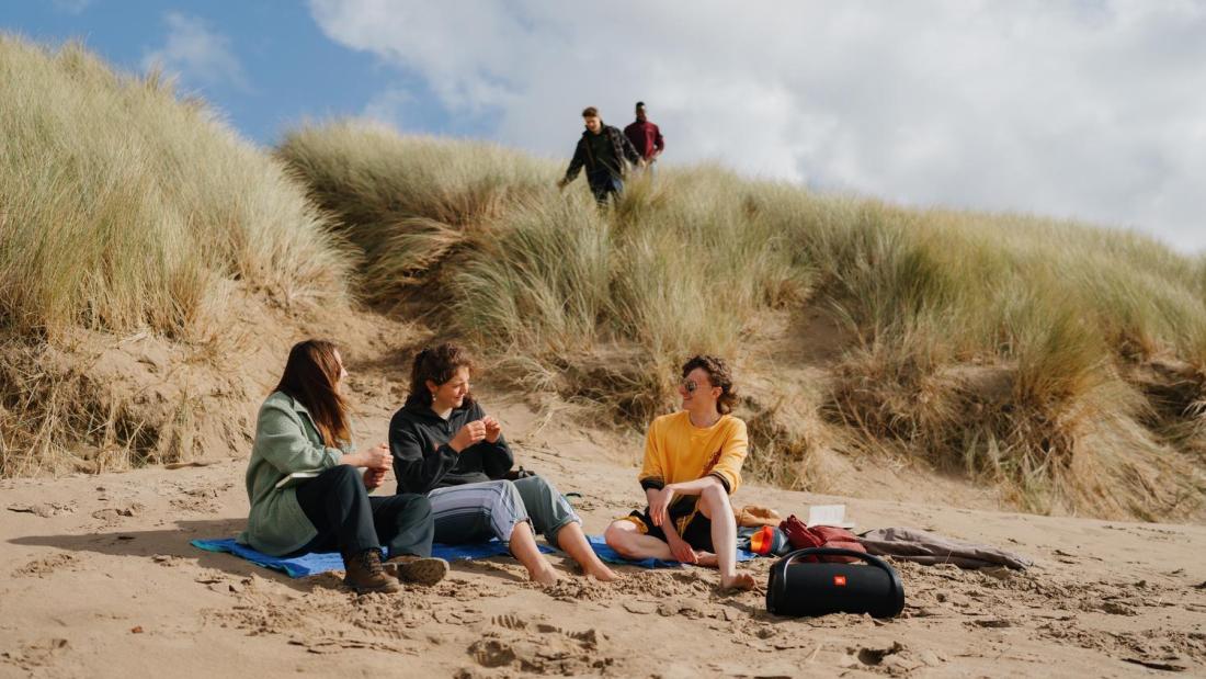 Three students sitting on the sand chatting and two students are walking towards them through the dunes behind them