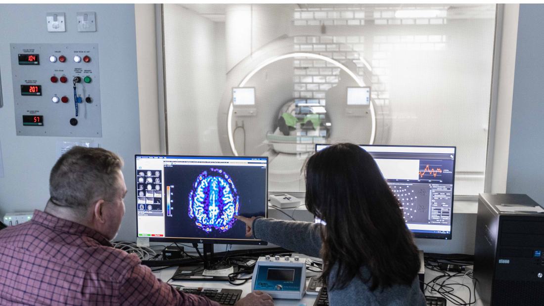 Two researchers discussing images of a brain on a computer screen, with an MRI scanner in the background.