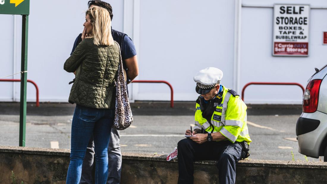 Police Officer sitting on a wall writing a speading fine whilst two adults ar standing