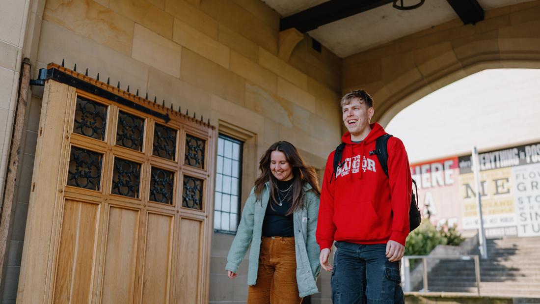 Two students walking through memorial arch one wearing a red Bangor University hoodie the other a blue coat